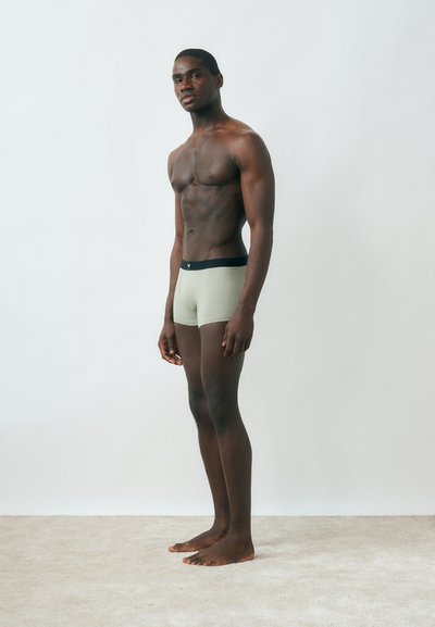 Γυμνός άντρας όρθιος ξυπόλητος σε χαλί, φορώντας ανοιχτόχρωμα εσώρουχα boxer με σκούρο ζωνάρι, μπροστά σε απλό ανοιχτό φόντο.