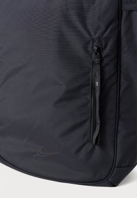 Artimiausias juodo audinio maišas su vertikaliu užtrauktuku ir mažai iškilusia Nike swoosh logotipo detalė apačios kampe.