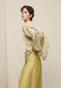 Femme aux cheveux courts et bouclés portant un chemisier transparent à motif papillon et un pantalon jaune taille haute à jambes larges, posant de profil avec les mains sur les hanches.