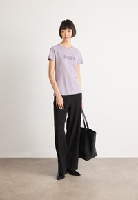 Moteris stovi laikydama juodą rankinę, vilkėdama levandų spalvos "BOSS" marškinėlį, juodas plačias kelnes, juodus batus ir sidabrinę apyrankę, neutralus fonas.