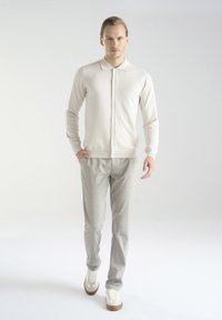 Jasnobeżowy kardigan z guzikami, zestawiony z jasnoszarymi spodniami w kratkę i casualowymi białymi trampkami z brązowymi podeszwami. Gładka tekstura.