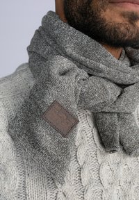 Bufanda de punto gris con un patrón texturizado y una etiqueta de cuero marrón. La bufanda está envuelta de manera suelta alrededor de un suéter de punto trenzado de color claro.