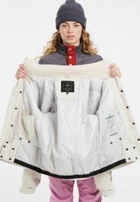 Giacca bianca trapuntata con rifiniture in velluto a coste, colletto morbido e molteplici tasche interne. Etichetta visibile all'interno. Modello che indossa un cappello grigio e un maglione.