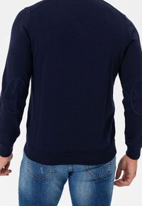 Suéter azul marino con dobladillo y puños de canalé. Con parches de ante en los codos del mismo color. Llevado sobre vaqueros azul claro.