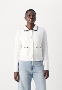 Biely cardigan s čiernym lemovaním goliera, zlatými gombíkmi a dvoma prednými vreckami. Vyrobený z jemného materiálu s rebrovanými manžetami a lemom.