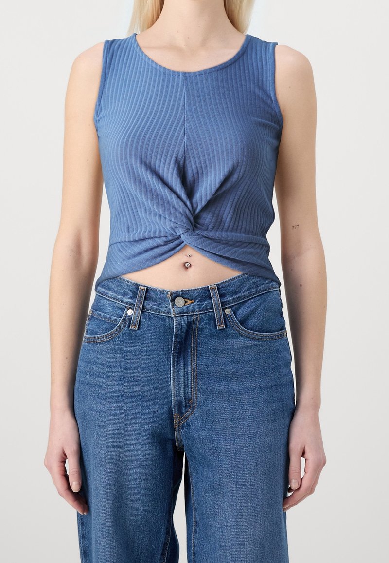 Femme portant un crop top bleu côtelé sans manches torsadé à la taille, associé à un jean bleu taille haute et des piercings au nombril visibles.