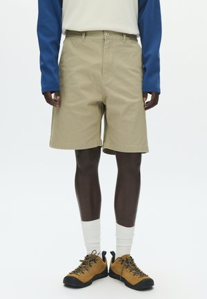 Béžové bavlnené šortky s voľným strihom, vybavené zapínaním na gombík, bočnými vreckami a hladkou textúrou. Nosené s tmavými topánkami a bielymi ponožkami.