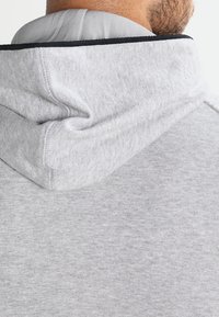 Světle šedá mikina s kapucí, která vykazuje měkkou texturu, ribbed výstřih a viditelné švy na kapuci, což zdůrazňuje její ležérní design.