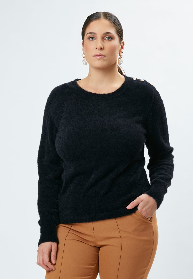 Sort, fluffy sweater med rund hals, lange ærmer og dekorative guldknapper på skulderen, kombineret med tætsiddende bukser i tan.