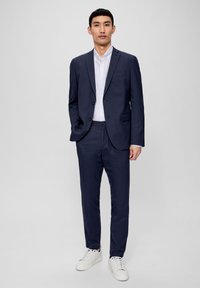 Dunkelblauer Anzug mit Reverskragen, zwei Knöpfen und schmal geschnittenen Hosen, kombiniert mit einem weißen Hemd und weißen Sneakers.