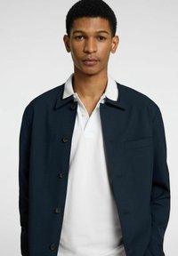 Veste bleu marine avec un col blanc, dotée de boutons et d'une poche. Portée par-dessus un polo blanc, mettant en valeur une texture lisse.