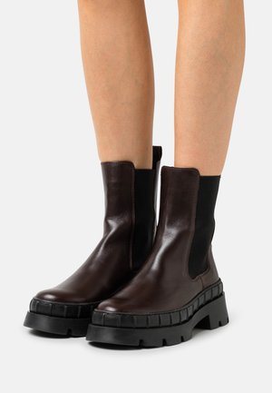 Bottines en cuir marron avec des panneaux élastiques sur les côtés et une semelle épaisse noire à texture striée. Fini lisse et design minimaliste.