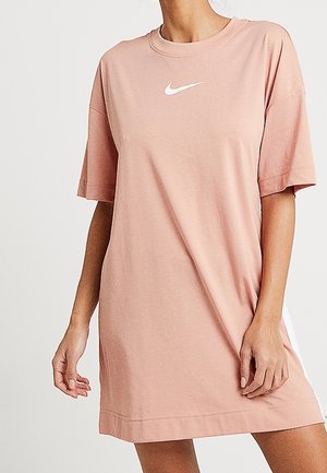 Donna che indossa un vestito rosa Nike, corto e a maniche corte, con logo swoosh bianco, in piedi contro uno sfondo chiaro e semplice.