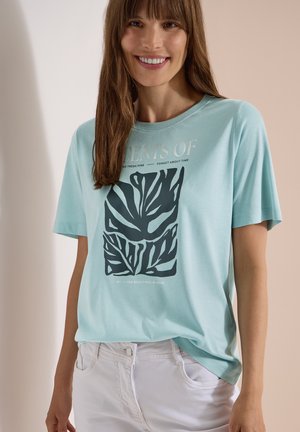 Femme souriante portant un t-shirt bleu clair avec un motif de feuilles foncées et un texte, associée à un pantalon blanc, debout devant un fond beige.