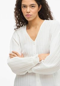 Hvid bluse med V-hals, teksturerede vertikale striber og puffede ærmer med elastiske manchetter. Har knapper foran.