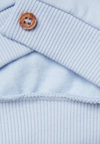 Světle modrá bavlněná tkanina s vroubkovanou strukturou, opatřená hnědým dřevěným knoflíkem a hladkou vnitřní vrstvou.