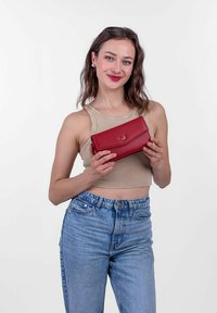 Červená kožená peněženka s hladkým povrchem, nabízí skládací design a vyražené logo na přední straně. Model ji drží proti jednoduchému pozadí.