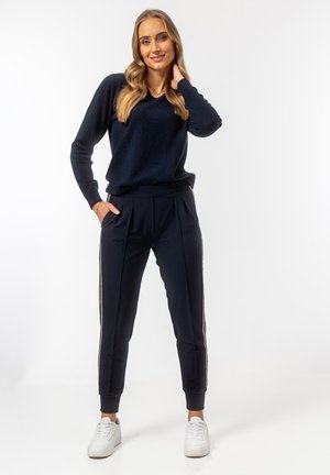 Granatowy sweter dzianinowy z dekoltem w serek, zestawiony z granatowymi spodniami plisowanymi z bocznymi paskami, uzupełniony białymi sneakersami.