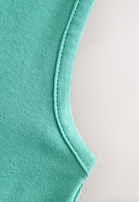 Närbild av en teal-färgad tygärm med synlig sömnad och ett mjukt, strukturerat stickmönster på en ljus bakgrund.