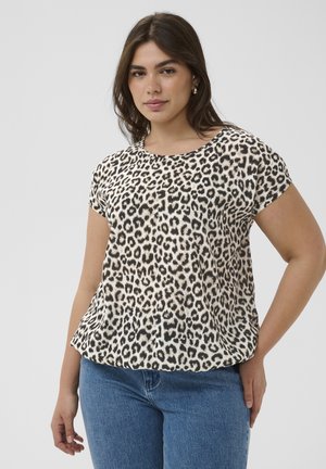 Woman wearing a short-sleeve leopard print top and blue jeans, standing with one hand on her hip against a plain background.