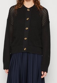 Svart stickad cardigan med öppen väv, ribbat nederkant och sex stora bruna knappar. Långa ärmar med krage.