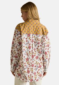 Naine, kellel on blonde juuksed, kannab pikka varrukatega lillelist särki, millel on valge põhi ja sinepikollane kaelus, millel on punased, sinised ja rohelised lillemuster.