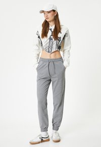 Korte sweatshirt met een witte body en zwart grafisch ontwerp, gecombineerd met grijze joggers. Het model draagt een witte pet en sneakers.