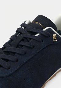 Ναυτικό σουέντ αθλητικό παπούτσι με υφή στην επιφάνεια, λευκά κορδόνια, χρυσές λεπτομέρειες λογότυπου και σόλα από καουτσούκ χρώματος γκαμάν. Τρύπες στα πλάγια για αερισμό.