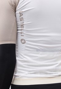 Εγγύς πλάνο ενός ατόμου που φοράει λευκή και μπεζ φανέλα ποδηλασίας με το λογότυπο "CASTELLI" και μαύρο μανίκι στο χέρι.