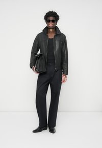 Black leather jacket with high collar, paired with a black fitted tank top, tailored black trousers, and pointed black shoes; holding a bag.