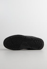 Semelle en caoutchouc noir d'une basket avec un motif de bande de roulement en zigzag et le logo FILA embossé au centre, présenté sur une surface unie.