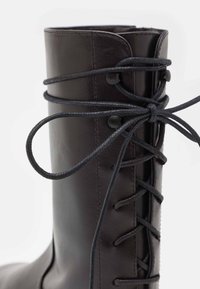 Bottine en cuir noir avec un design à lacets, texture lisse, œillets renforcés et détails tressés sur le côté.