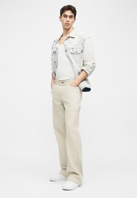 Jeune homme debout, mains dans les poches, portant une veste en denim clair à motifs, un débardeur blanc, un pantalon en velours côtelé beige et des baskets blanches.