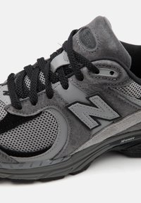 Šedá sportovní bota s materiály z méně a semiše, štíhlý design, vrstvené textury, polstrovaný límec a výrazné logo na boku.