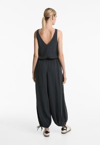 Temno siva brezrokavna kombinezonska obleka z V-izrezom na hrbtu, sproščenega kroja, zrahljano pasom in širokimi, zoženimi nogami. Ima poudarke s čim na robovih.