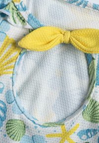Tissu coloré avec des motifs de coquillages et d'étoiles de mer bleu clair, jaune et vert. Comprend un accent en nœud jaune et un détail en découpe circulaire.