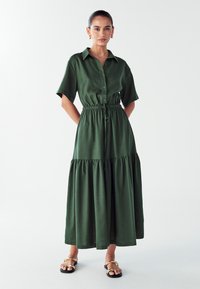 Zielona, krótko rękawowa sukienka na guziki z elastycznym pasem i warstwowym dołem. Materiał wydaje się miękki o gładkiej teksturze. Noszona z sandałami.