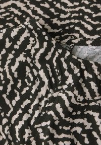Schwarzer und beiger Stoff mit einem gewellten, abstrakten Muster. Das Material ist leicht und hat eine weiche Textur, mit sichtbaren Stichdetails entlang des Randes.