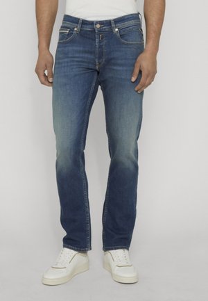 Mann, der blaue ausgewaschene Jeans und weiße Turnschuhe trägt, steht vor einem schlichten Hintergrund.