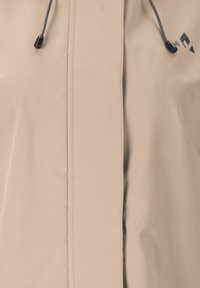 Beige jacka med en slät textur, framdragkedja, justerbar dragsko och en svart logotypaccent nära kragen.