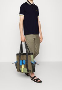 Sac fourre-tout rectangulaire multicolore en vert, bleu et jaune avec une surface texturée, des sangles noires et de multiples poches extérieures.