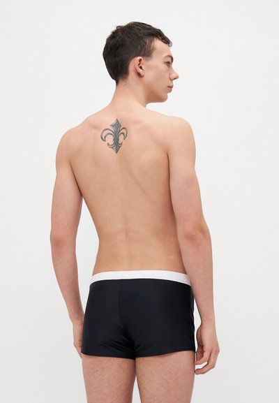 Un short de bain noir avec une taille blanche, design ajusté et texture lisse, vu de dos. Un tatouage de fleur de lys est visible.
