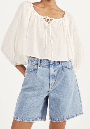 Persoon die een witte blouse met lange mouwen en een strik aan de voorkant draagt, met lichtblauwe, hooggetailleerde denim shorts met een ruwe zoom.