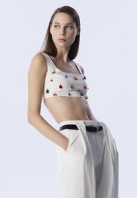 Witte crop top met kleurrijke decoratieve kralen. Het model draagt losse witte broeken met een zwarte riem, wat een moderne, high-fashion uitstraling geeft.