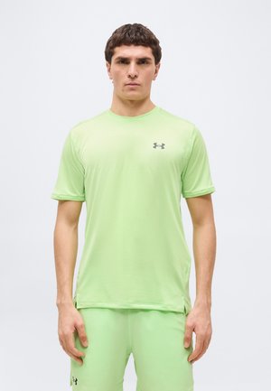 Muž v svetlozelenej krátkosti Under Armour a zodpovedajúcich šortkách stojaci pred jednoduchým bielym pozadím.