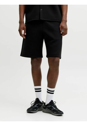 Person steht und trägt schwarze Shorts, weiße Socken mit zwei schwarzen Streifen und schwarze Sportschuhe vor einem schlichten hellen Hintergrund.
