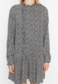 Schwarz-weiß gemustertes Kleid mit einem gerafften Ausschnitt und langen Ärmeln, mit einem gestuften Saum und geometrischen Designs auf dem gesamten Kleid.