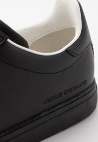 Sneaker en cuir noir avec des accents blancs, présentant un bout arrondi, des lignes épurées, des détails cousus et un logo "Armani Exchange" en relief.