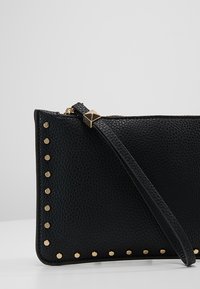 Pochette bracelet noire texturée avec des clous dorés le long des bords et une bandoulière noire avec un ornement doré en forme de pyramide près de la fermeture éclair.