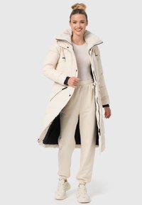 Lång krämfärgad pufferjacka med hög krage, dragkedja och knäppning, samt sidofickor, ihopparad med beige mjukisbyxor och ljusa sneakers.